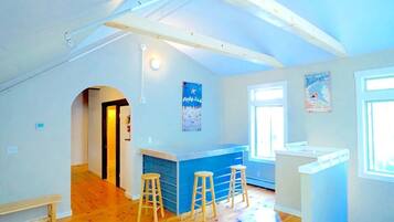 An open-plan living space with a kitchen area, wooden flooring, and a high ceiling with exposed beams.