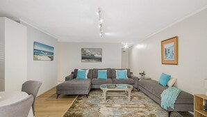 Una sala de estar con un sofá modular gris, una mesa de centro de vidrio y un tapete estampado. Hay dos cuadros en las paredes y una mesita auxiliar con una planta.