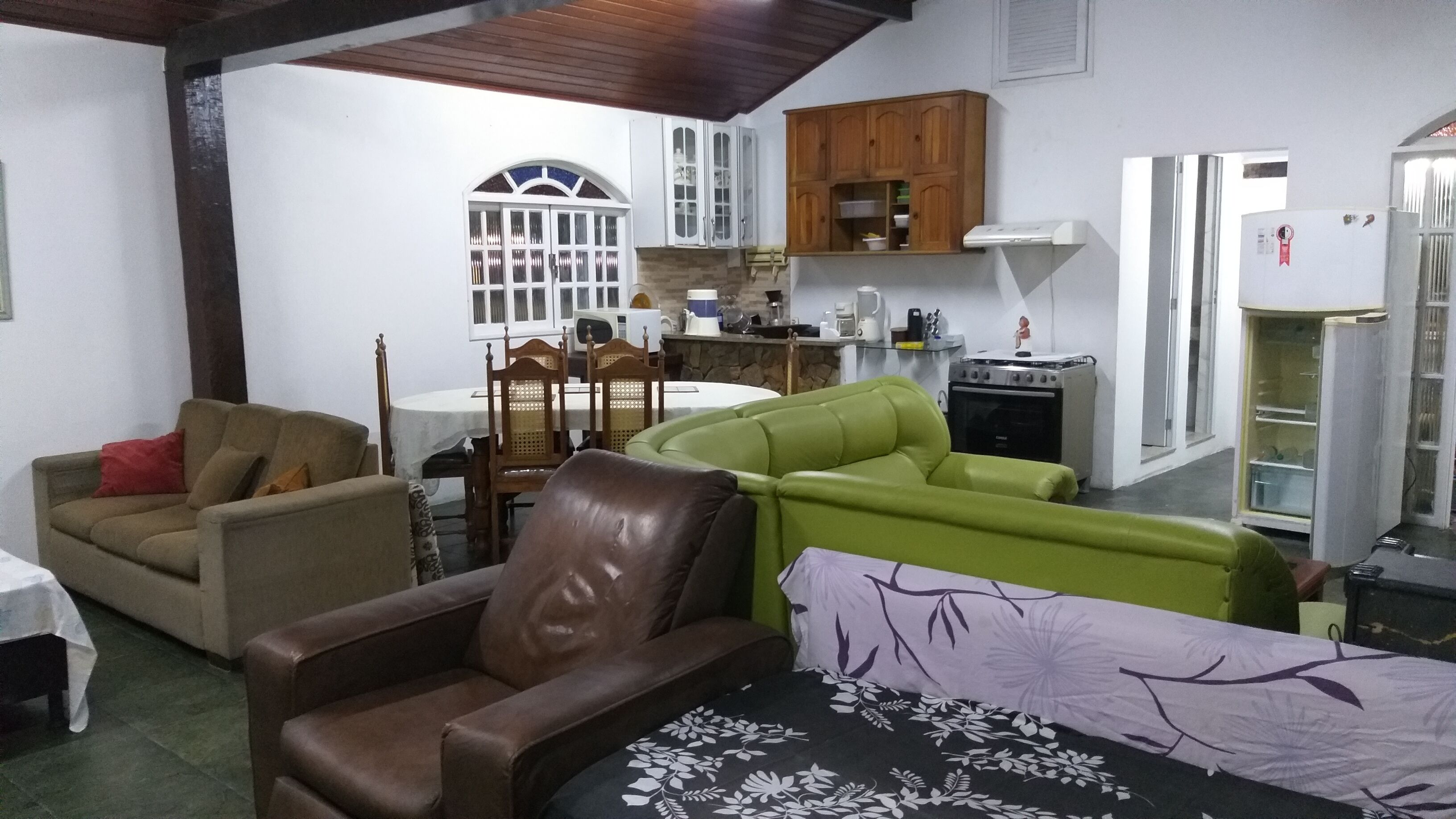 Een woonkamer met een bruine leren bank, een groene bank en een tafel met een tafelkleed met bloemen. Er is een houten eettafel met stoelen en een raam met een decoratieve boog. In de achtergrond is een keuken met houten kastjes en apparatuur zichtbaar.