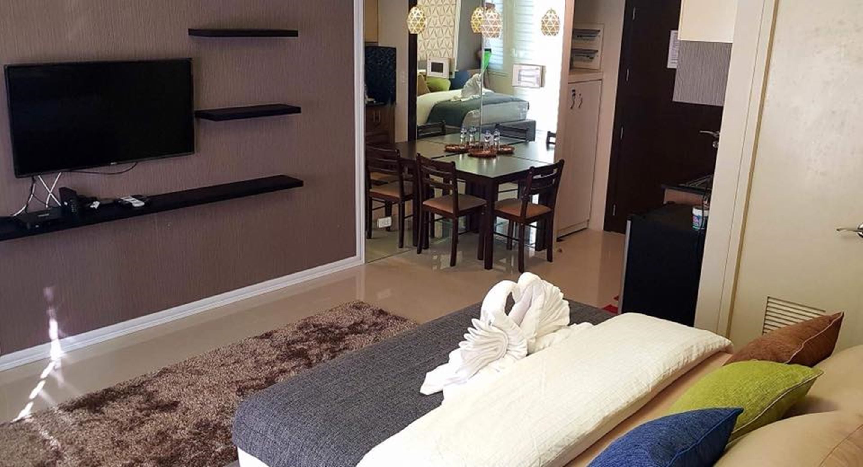 Een moderne hotelkamer met een flatscreen televisie aan de muur, een zithoek met een glazen tafel en stoelen, en een opgemaakt bed met een handdoek.