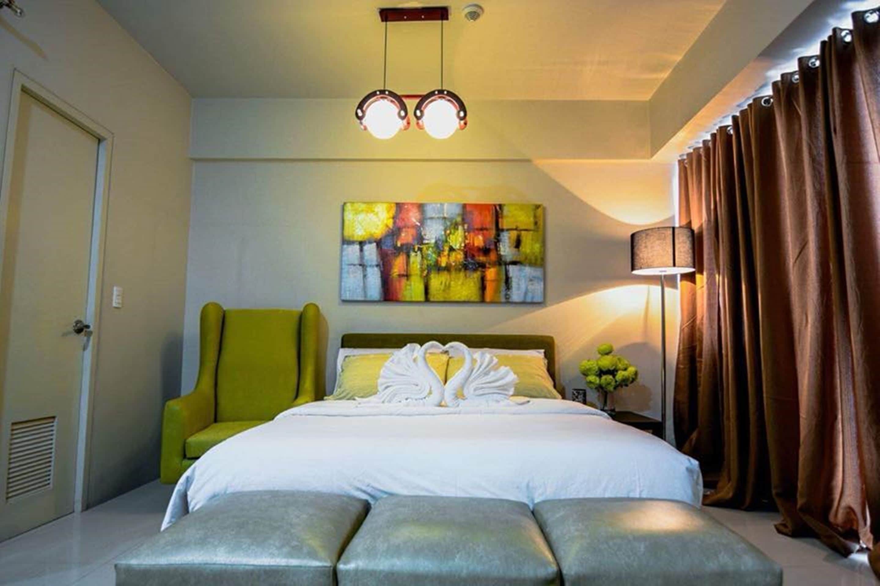 Una habitación de hotel con una cama, un sillón verde, un cuadro abstracto de colores, una lámpara y cortinas.