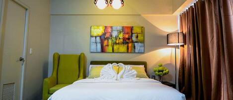 Una habitación de hotel con una cama, un sillón verde, un cuadro abstracto de colores, una lámpara y cortinas.