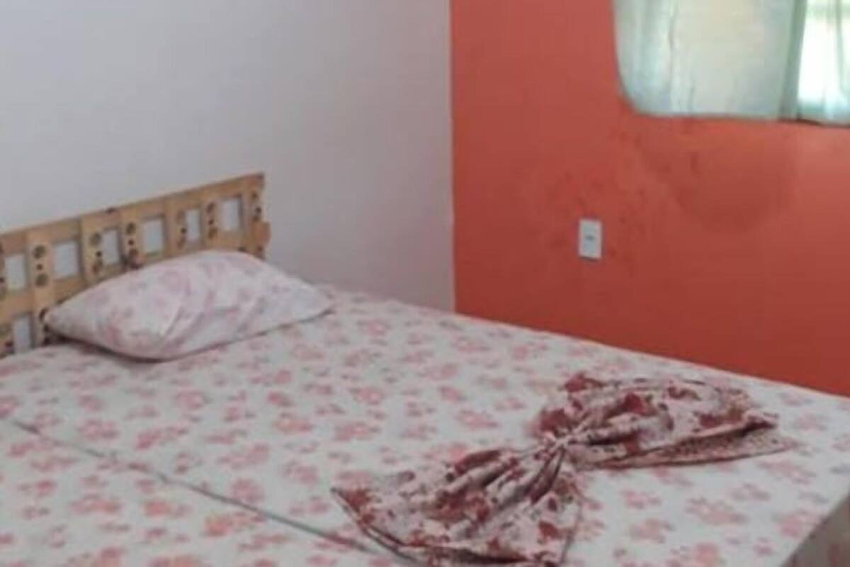 Un lit simple avec une couvre-lit à motifs floraux et une tête de lit en bois, dans une pièce dont le mur est orange et qui possède une fenêtre ornée d’un rideau léger.