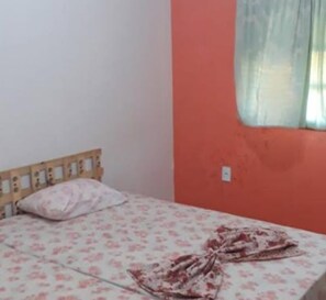 A single bed with a floral bedspread and a wooden headboard in a room with an orange wall and a window with a sheer curtain. - Pousada Villa Vittoria (Caucaia)