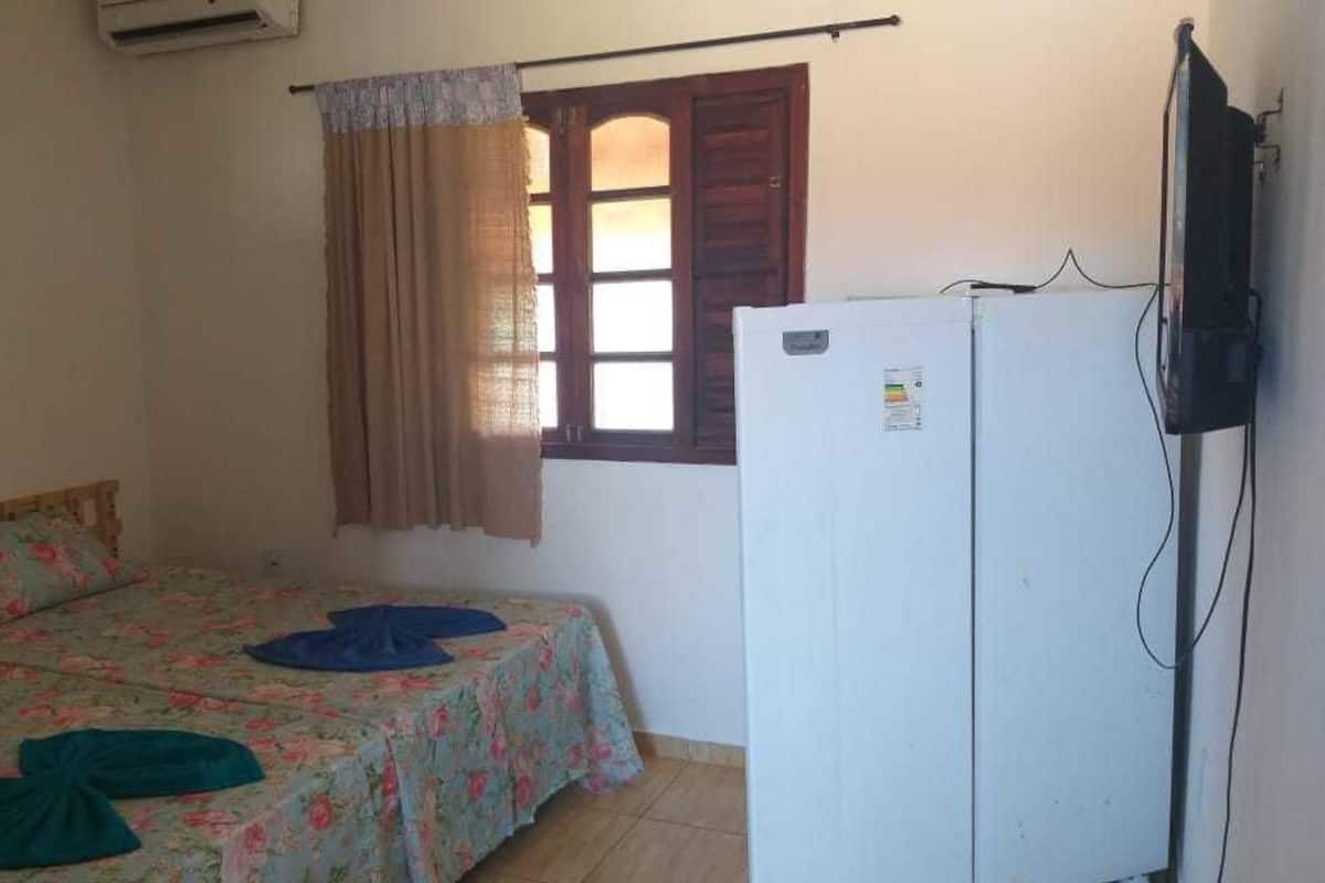 Une petite chambre avec un lit simple, un réfrigérateur blanc, une fenêtre avec des rideaux et un téléviseur fixé au mur.