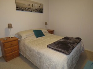 A neatly made bed with a quilted cover, two bedside tables with lamps, a small drawer, and a painting on the wall.