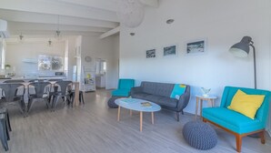 A modern living room with a grey sofa, teal armchairs, and a round ottoman. There is a dining area with a white table and chairs, and a kitchen in the background. - 63 Marlin by Smiths Beach (Smiths Beach)