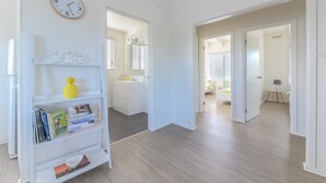 A spacious room with a wooden floor, a white wall clock, and a bookshelf with books and a pineapple. - 63 Marlin by Smiths Beach (Smiths Beach)