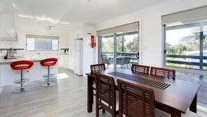 A modern kitchen with white cabinets, a stainless steel refrigerator, and a red fire extinguisher. There is a dining area with a wooden table and chairs, and large windows offering a view of the outdoors. - 47 Anderson with Pool in Cowes (Cowes)