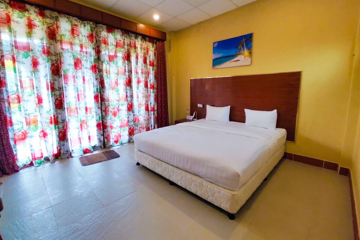 A hotel room with a large bed, floral curtains, a wooden headboard, and a painting of a beach scene.