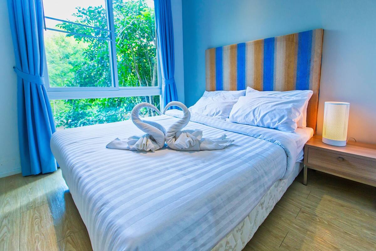 Una cama bien hecha con sábanas blancas y dos toallas con forma de cisne, un cabecero de madera, una mesita de noche con lámpara y una ventana con cortinas azules.