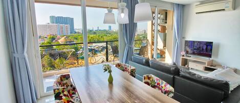 A modern living room with a wooden dining table, colorful chairs, a black sofa, a TV, and a balcony with a city view.