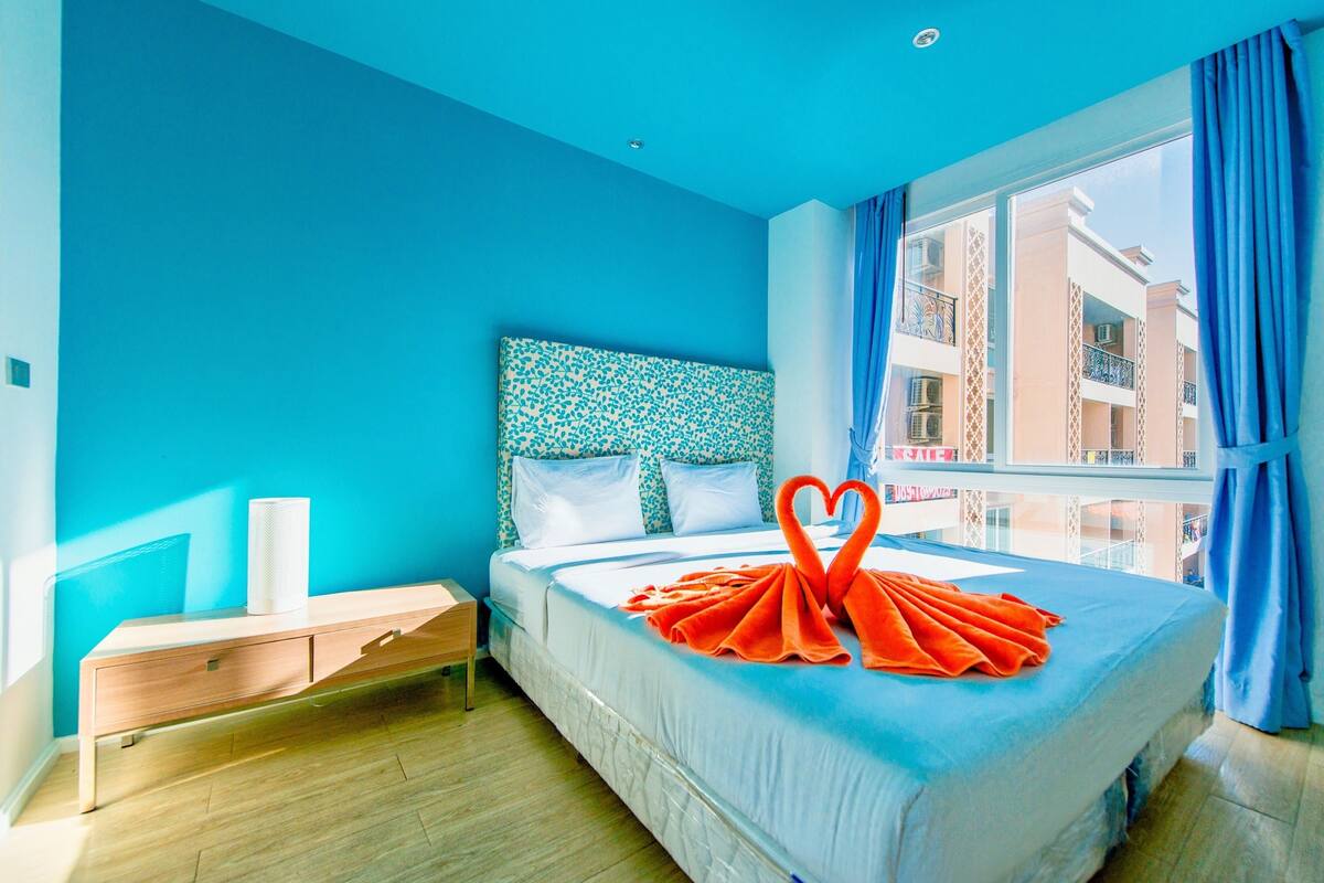 Habitación de hotel con cama, mesita de noche, ventana con vista a edificios y pared azul.