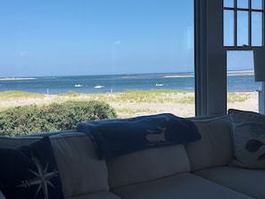 Vista desde una ventana con sofá y cojines, hacia una playa con barcos y cielo despejado.