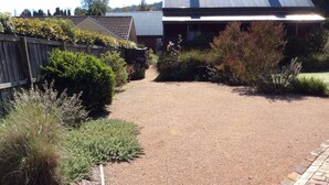 A gravel driveway leading to a house with a metal roof, surrounded by various shrubs and a wooden fence. - Studio Rouge in heart of old Bowral only minutes walk to shops and restaurants (Bowral)