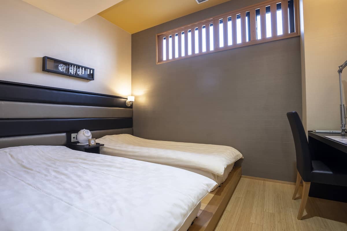 A hotel room with two single beds, a desk with a chair, a wall clock, and a window with blinds.