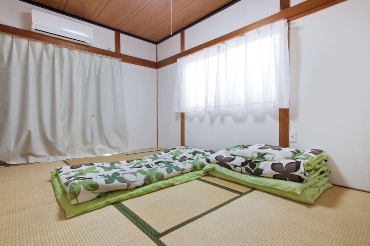 A room with tatami flooring, a bed with green and brown floral bedding, a window with sheer curtains, and a wall-mounted air conditioner.