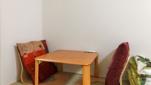 A small wooden table with a remote control on it, placed in a corner of a room with a red cushion and a green mat on the floor. - Guesthouse SAYURI 2 (Osaka)