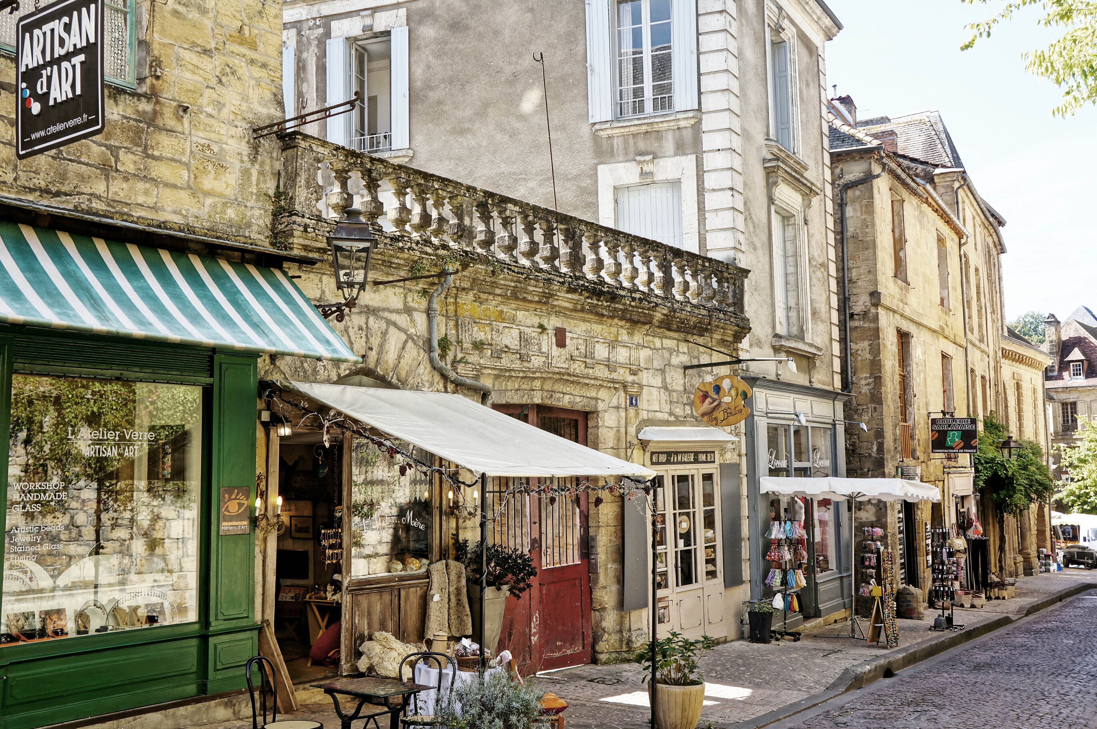 Une rue commerçante, avec notamment une boutique dont la devanture est bien visible et qui affiche une enseigne indiquant « Artisan d’Art ».