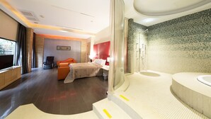 A modern hotel room with a large bed, a seating area with a sofa and armchairs, a separate bathroom with a bathtub and shower, and a large window providing a view of the outdoors.