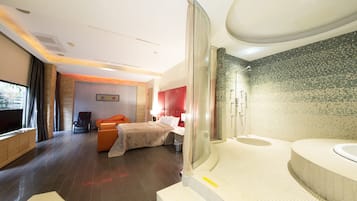 A modern hotel room with a large bed, a seating area with a sofa and armchairs, a separate bathroom with a bathtub and shower, and a large window providing a view of the outdoors.
