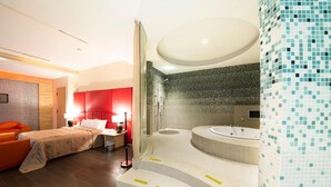 A modern hotel room with a bed, a red headboard, a shower area, and a large bathtub.