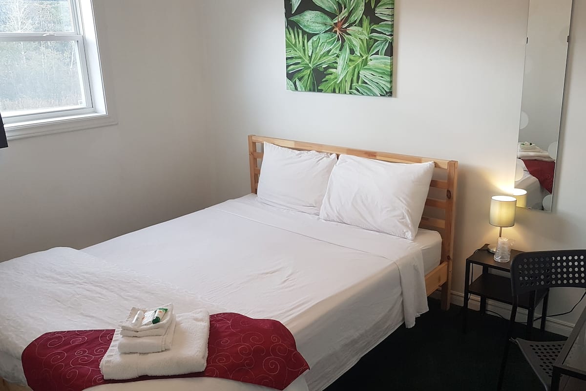 Una cama bien hecha con sábanas blancas, un cojín rojo y una toalla a un lado. Una mesita con una lámpara y una silla están junto a la cama. Un cuadro de hojas verdes adorna la pared sobre la cama.