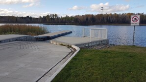 Un muelle de hormigón se adentra en un lago tranquilo, rodeado de zonas verdes y una orilla boscosa.