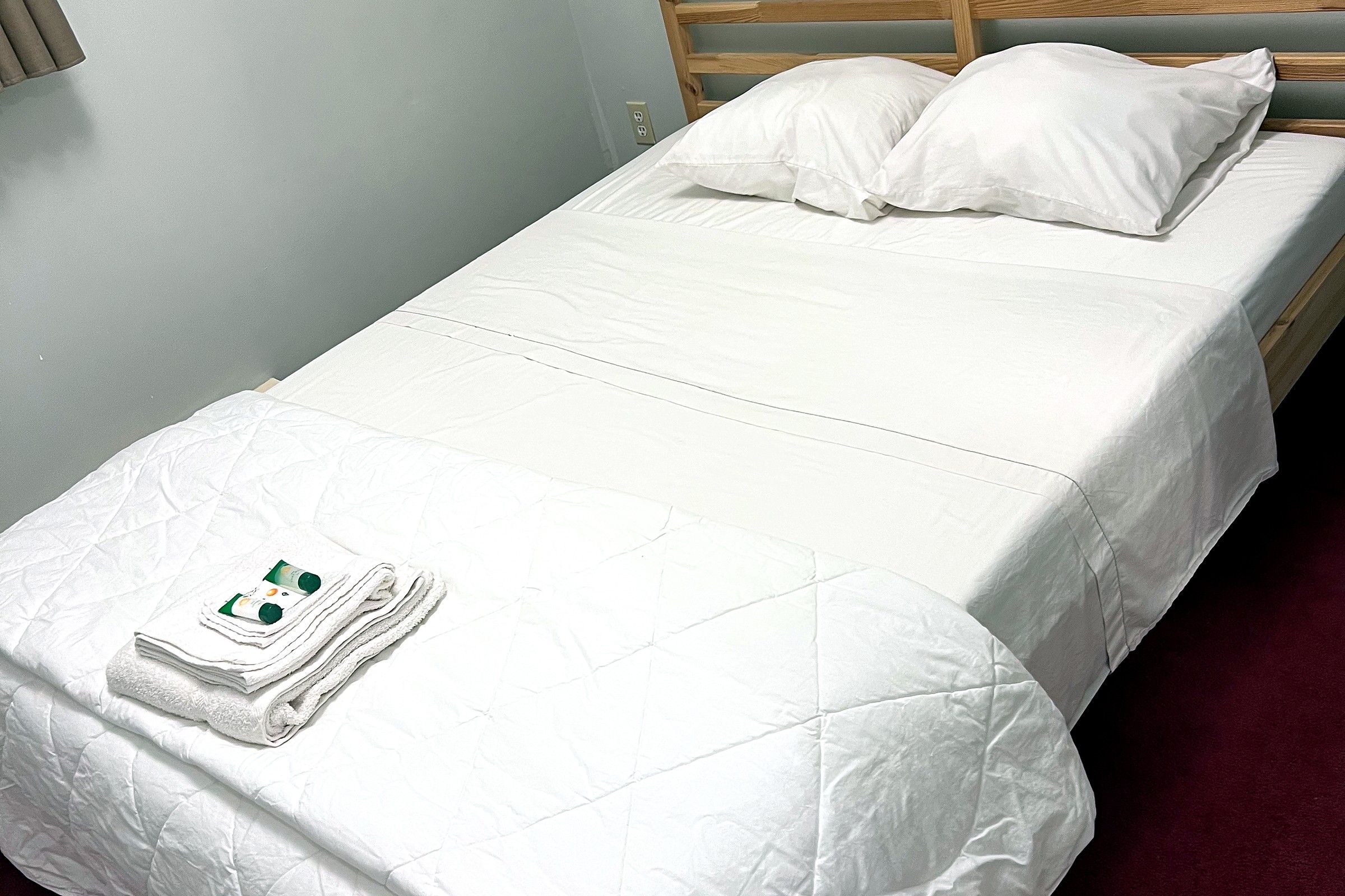 Una cama individual con ropa blanca y almohadas, una toalla doblada y una botella de loción sobre la cama.
