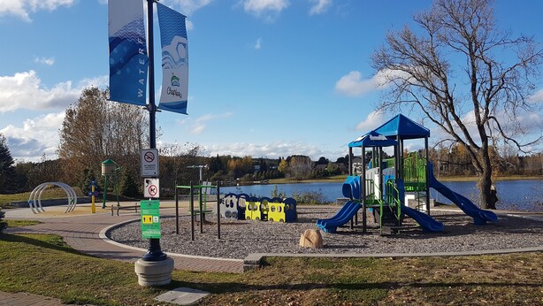 Un parque infantil con toboganes y estructuras para escalar cerca de un lago, rodeado de árboles y un cielo despejado.