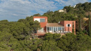 Une maison moderne à la façade rouge, dotée de grandes fenêtres et d’un patio entouré d’une végétation luxuriante.