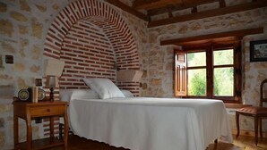 Een slaapkamer met een bakstenen boog, houten vloer, een bed met wit beddengoed, een nachtkastje met een lamp en een raam met uitzicht op groen.