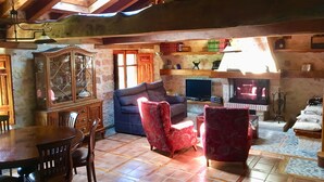 Een landelijke woonkamer met een houten plafond, een eettafel met stoelen, een bank, een rode fauteuil, een televisie en een open haard.