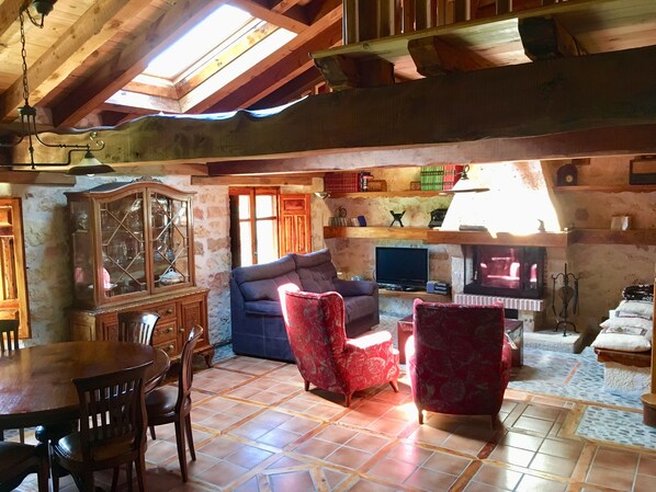 Un salon de style rustique avec un plafond en bois, une table à manger avec des chaises, un canapé, un fauteuil rouge, un téléviseur et une cheminée.