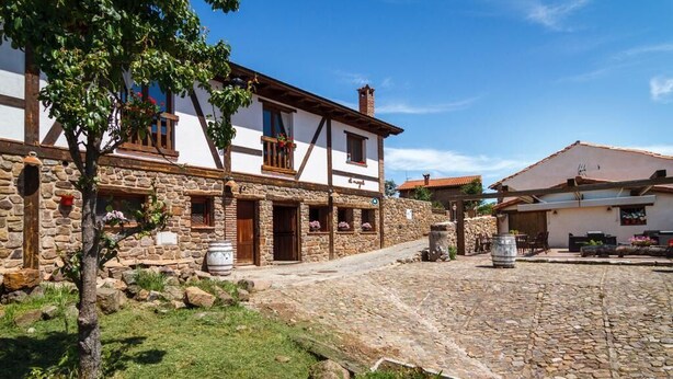 Un edificio in pietra tradizionale con facciata bianca e dettagli in legno, caratterizzato da un cortile in pietra e un piccolo albero in primo piano.