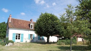 Una casa bianca con persiane blu e tetto in tegole, circondata da un prato e alberi.