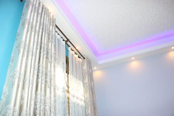 Una habitación con un amplio ventanal cubierto por cortinas de gasa con estampado de árboles, techo de vigas de madera con iluminación empotrada y una pared lisa.