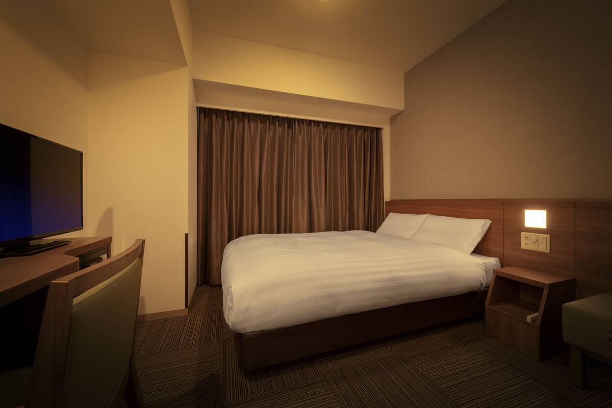 Una habitación de hotel con una cama, un escritorio con televisor, una mesita de noche y una lámpara en la pared.