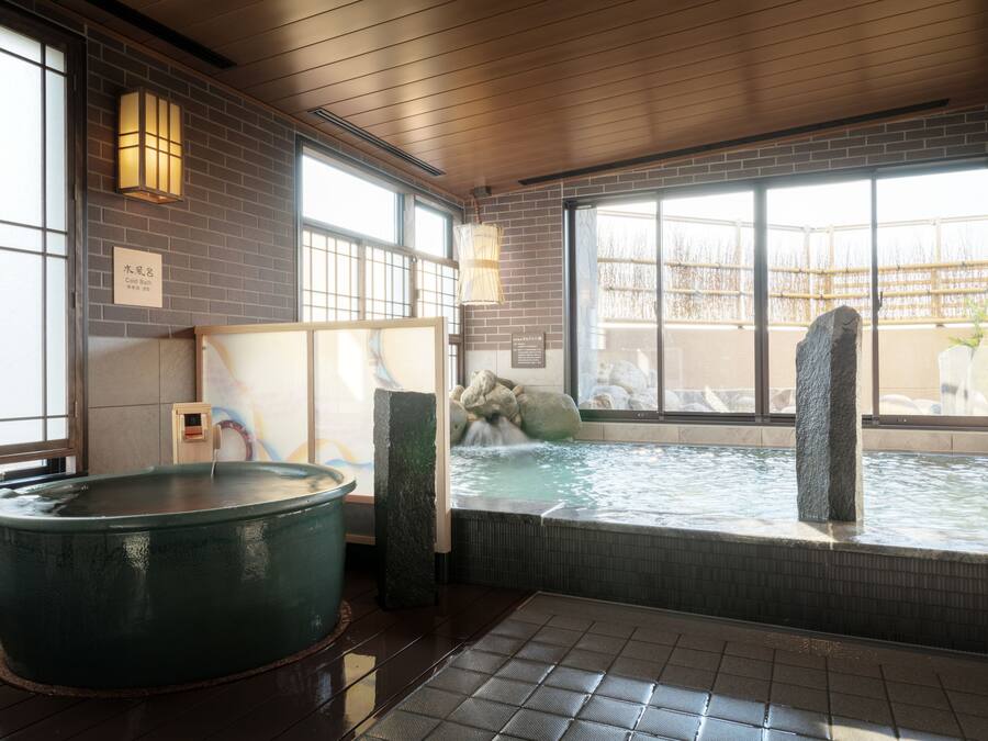 A traditional Japanese onsen with a large hot spring, wooden flooring, and a view of the outside through large windows.