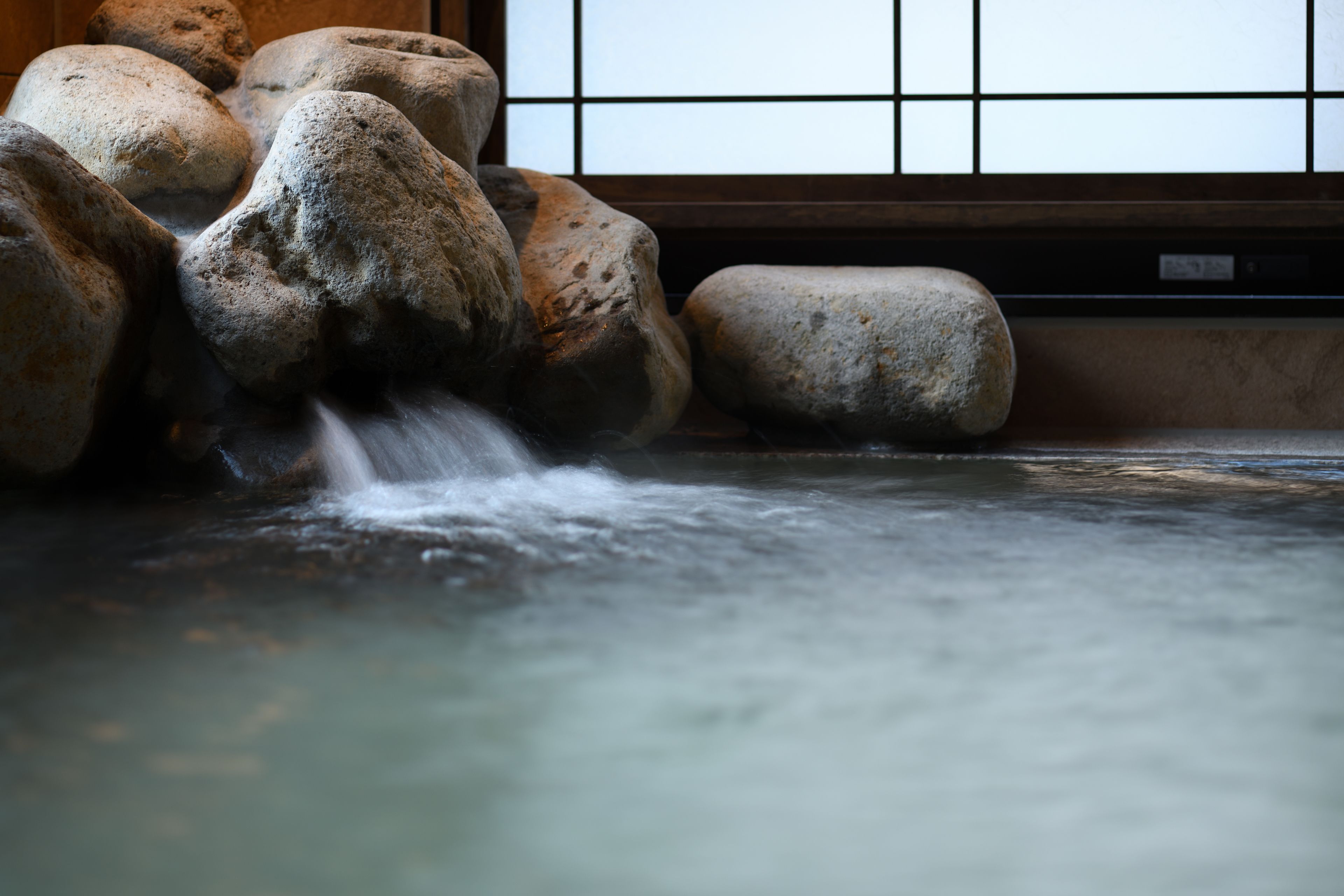 Un onsen japonés tradicional con una bañera de piedra rodeada de rocas y una ventana de madera con entramado.