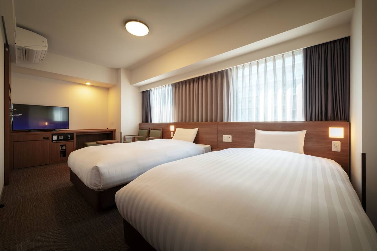 Una habitación de hotel con dos camas, televisión, escritorio y ventana con cortinas.