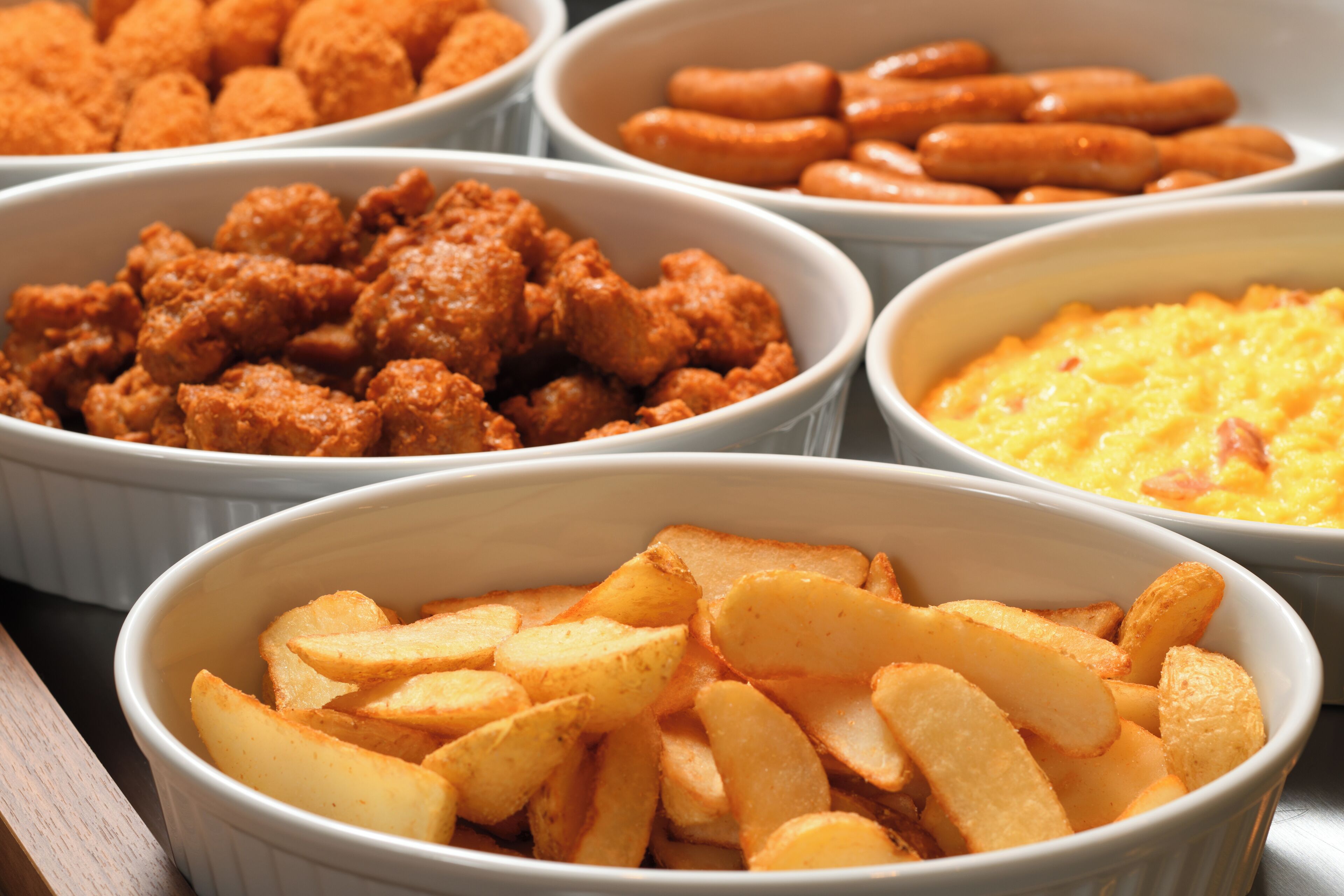 A variety of food items in white bowls, including what appears to be fried chicken, sausages, and potato wedges.