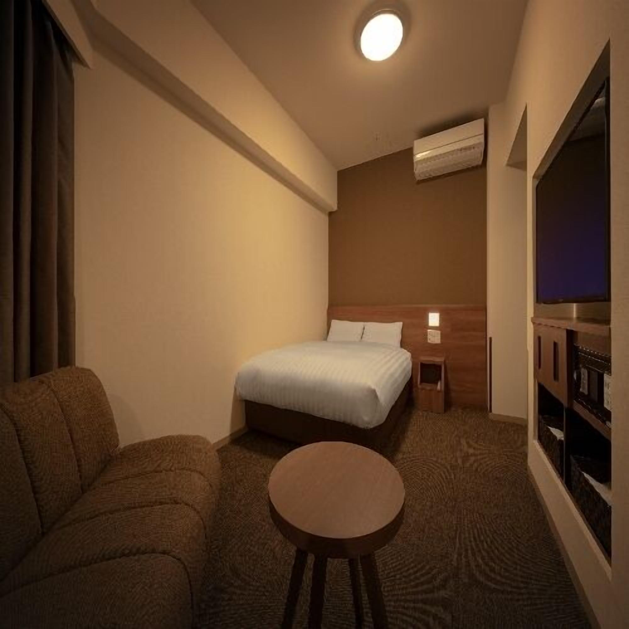 A compact hotel room with a bed, a small round table, a sofa, and a television.