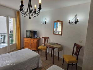 Une chambre d’hôtel comprenant un lit, une télévision, une armoire en bois, une table avec des chaises, un lustre et une fenêtre avec des rideaux.