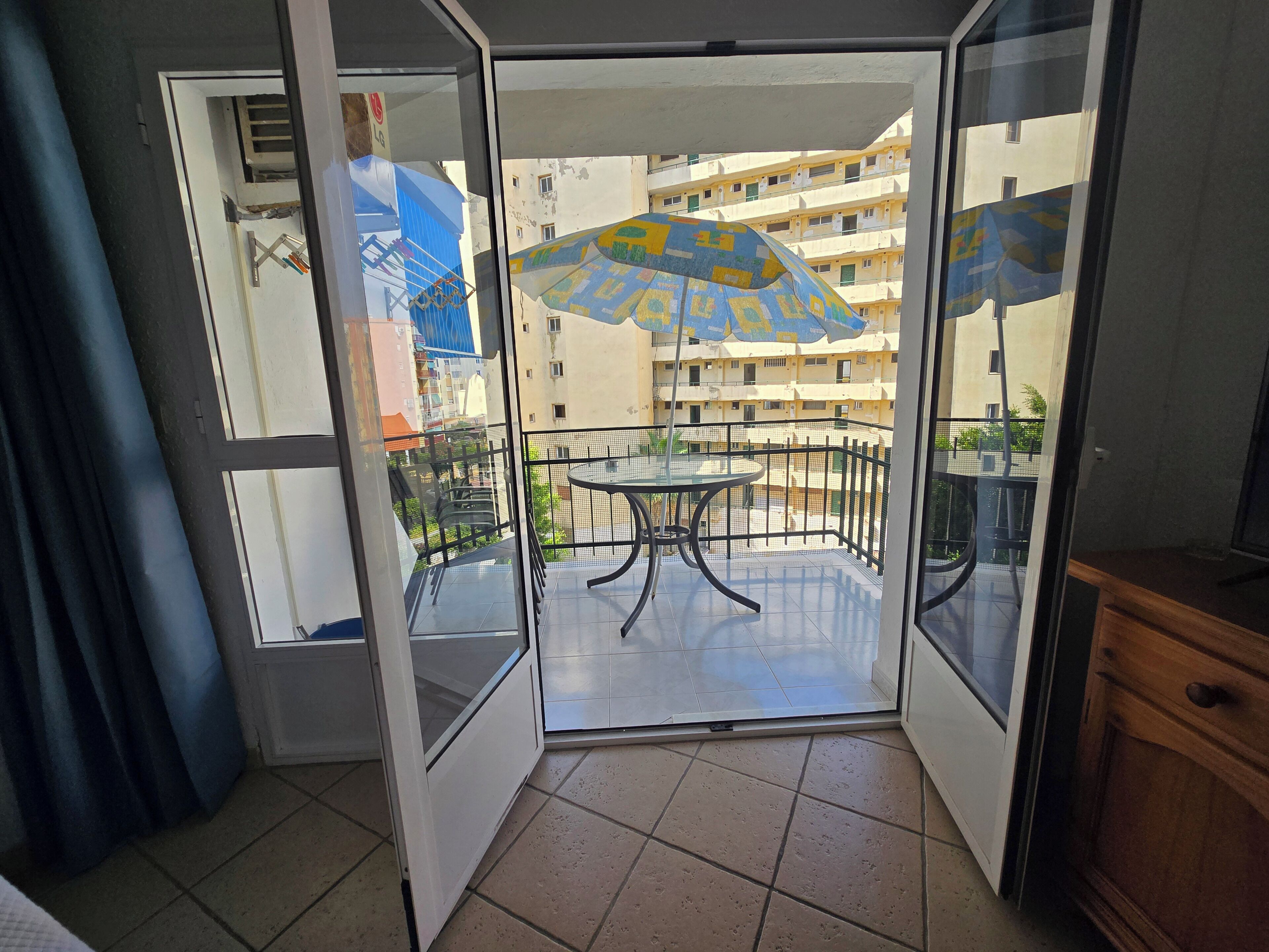 Een balkon met een tafel en stoelen, een blauwe en gele parasol, en uitzicht op een meerlaags gebouw.