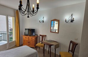Ett hotellrum med en säng, en TV, ett träskåp, ett bord med stolar, en kristallkrona och ett fönster med gardiner.