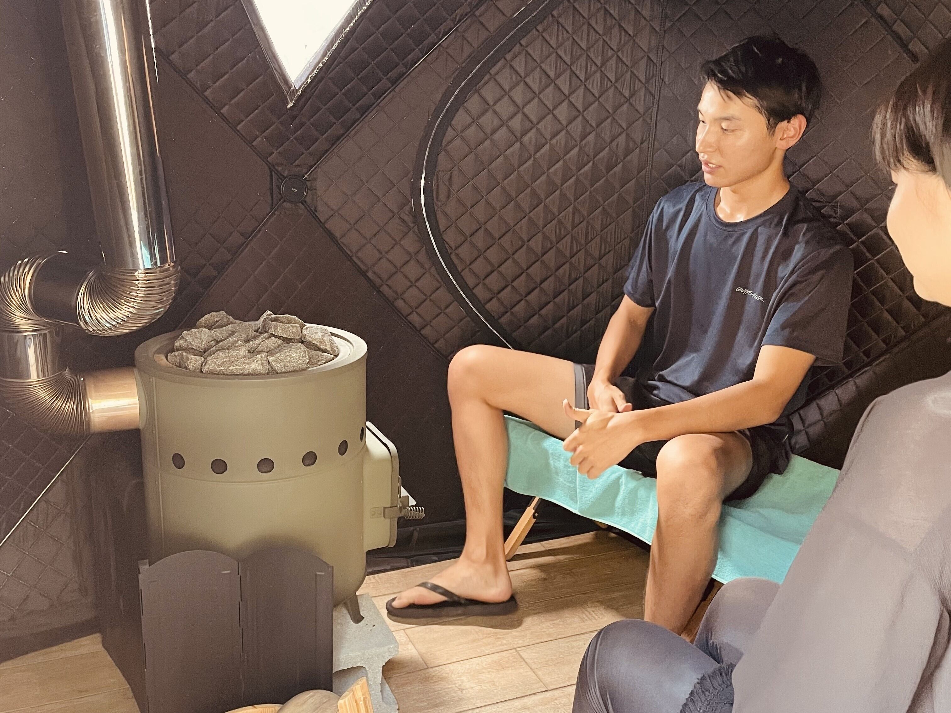 A room with a quilted wall, a small heater with rocks, and a person sitting on a bench.
