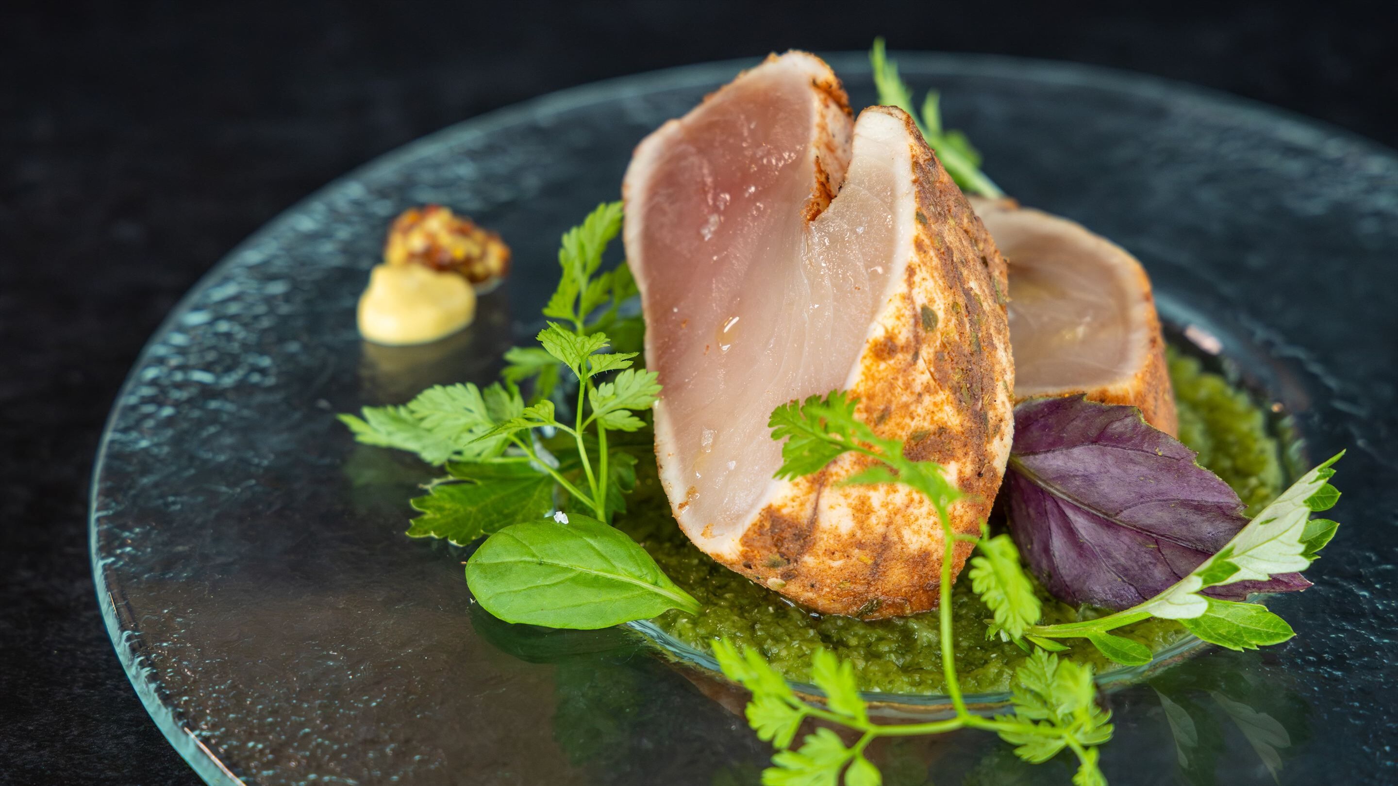 A dish with a piece of meat, greens, and a sauce on a glass plate.