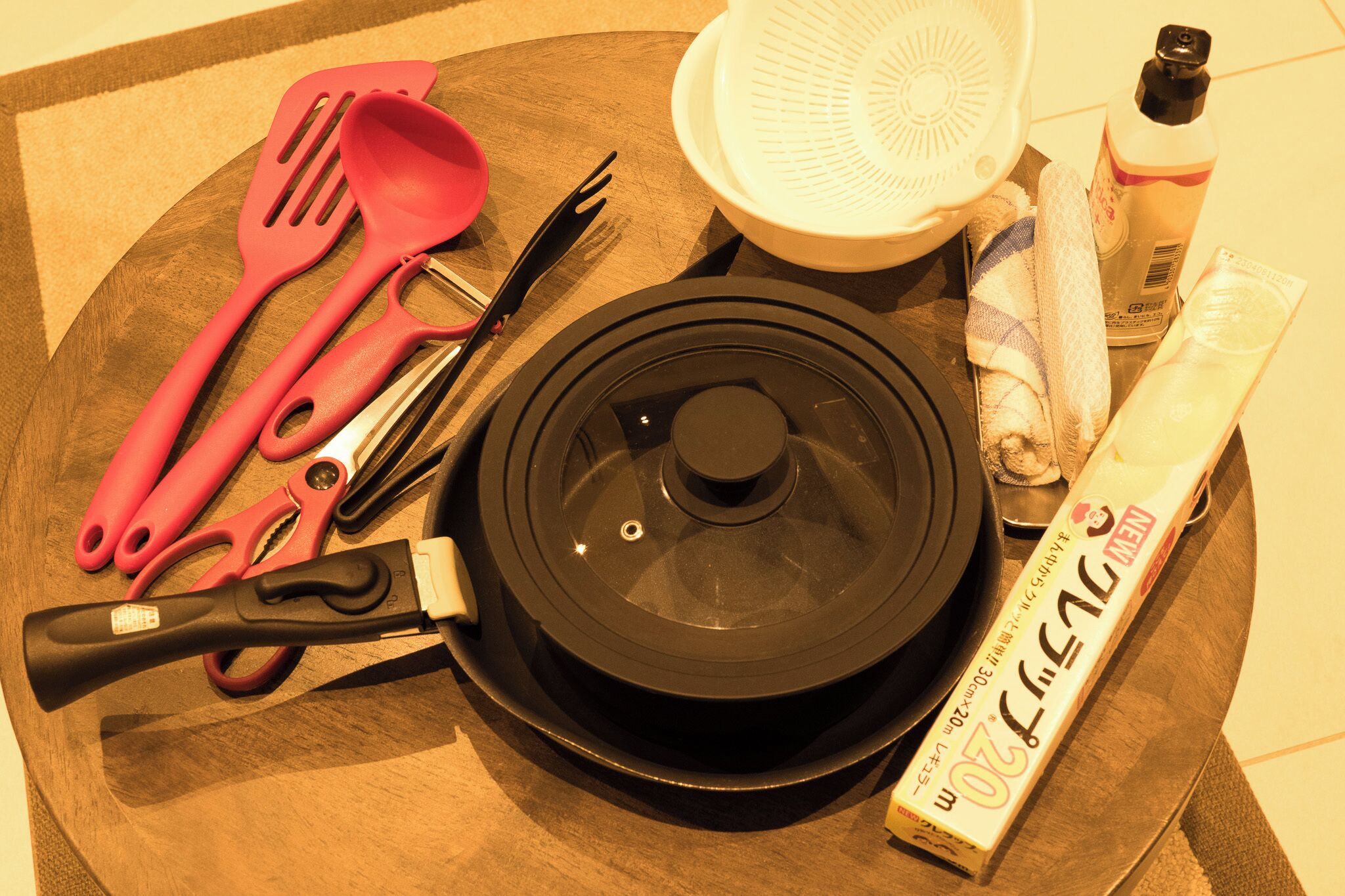 Una encimera de cocina con una olla negra, espátulas rosas, un colador blanco, una botella de detergente, una toalla y un tubo de crema.