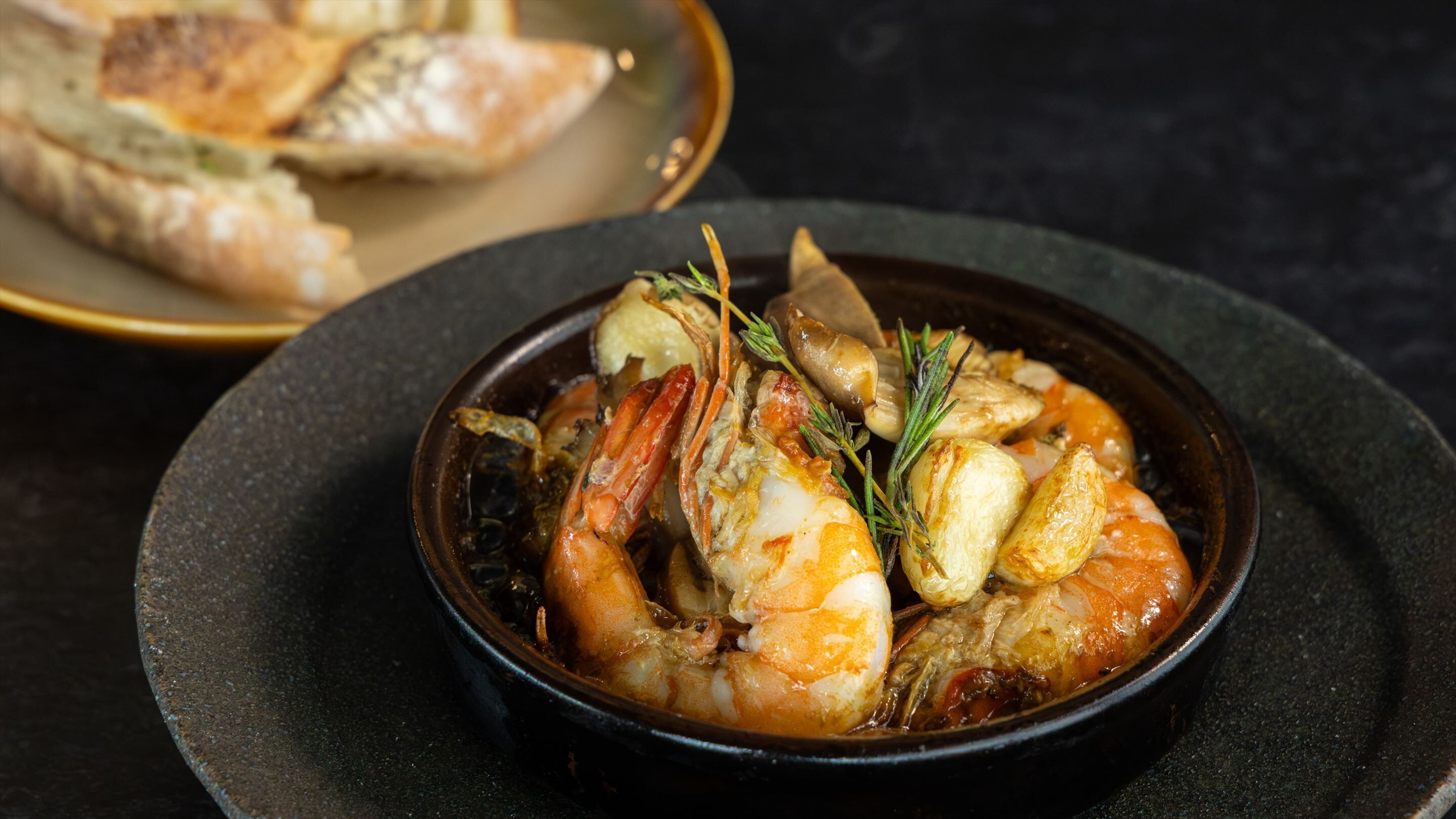 A bowl of seafood stew with shrimp and potatoes, garnished with herbs, served on a dark plate.